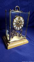 Front view of the Andrea by Sadek brass and glass mantle clock showing the Roman numeral dial and wooden base.