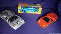 Three vintage battery-powered supercar toys displayed on purple surface: silver Lamborghini Countach model, red Hugo Boss Porsche 919 model, and boxed blue Computer Car.