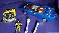 Photo showing blue 1984 Kenner Batmobile toy with orange tinted canopy, loose Batman figure, Joker and Batman PEZ dispensers, and packaged Batman figure in black and gold.