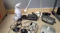 View of electronic items laid on floor including battery chargers, camera, lens, GPS, scale, and cables