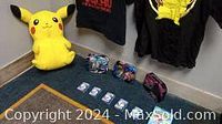 Photo showing large Pikachu plush doll, 3 Pokémon collector tins, and 2 black Pokémon-themed T-shirts hanging on the wall