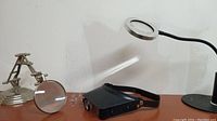 Magnifying light with goose neck, round lens, and round base on right; magnifying glass on movable metal stand in the middle; black plastic head magnifier in the foreground