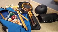 Photo showing tennis rackets, black carrying case, child's helmet, black curved protective gear, and croquet set inside blue bag.