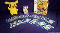 Photo showing a Pikachu plush doll, a deck box of Brilliant Stars Sword & Shield Pokemon trading cards, and a spread of approximately 40 cards face down with blue Pokeball backs, along with a Pokemon 500-piece Pikachu puzzle box.