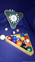 Full view of the pool ball clock on a triangular board with real pool balls arranged in a clock formation, wooden rack holding the balls, and several loose pool balls including the cue ball.