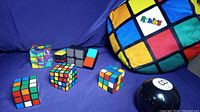 Wide view showing all five Rubik's cubes lined up with the Magic 8 Ball and the Rubik's Cube plush pillow on a blue fabric background.