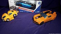 Lot view with yellow Bumblebee-style transformer toy car, orange transformer toy car with spoiler, and blue Gainer remote control car box in background.