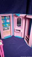 Open fold-out Barbie house showing illustrated bookshelf, TV, vanity with mirror, and closet printed inside with pink and teal colors.