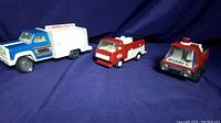 Three vintage metal toy trucks: Tonka Pepsi-Cola delivery truck, Tonka red fire truck, and Buddy L red fire truck on dark blue background.