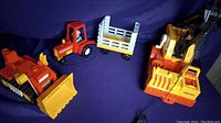Photo showing three vintage Fisher Price construction vehicles on a blue background: bulldozer, tractor with trailer, and crane.