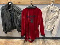 Three items hanging on hooks against a wooden wall: a black jacket, a red Playboy bathrobe, and a cream/off-white jacket.