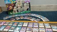 First photo shows two rows of Yu-Gi-Oh trading cards laid out on floor with Camelot 3D puzzle box behind them