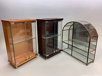 Three countertop display cabinets together; two wooden with glass doors and one arched mirrored brass with mirrored doors
