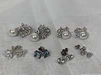 Overview of the six pairs of 925 sterling silver earrings with cubic zirconia stones and pearls on a white cloth surface.