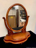Front and angled view of the antique hand carved walnut vanity shaving mirror showing the oval reflective surface, carved side supports with scroll details, and polished walnut base with curved edges.