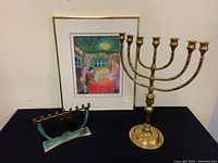 Photo showing framed signed print by Marianne M Vagler and two brass menorahs (one large, one smaller) on a black surface against white background.