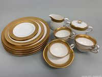 Full view of the dish set including plates stacked, teacups, saucers, creamer, and sugar bowl showing gold ornate detailing and white porcelain base.