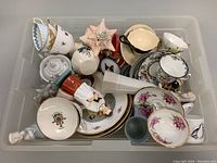 Top view of assorted chinaware including teacups, saucers, figurines, and a decorative sea shell all collected in a plastic bin.