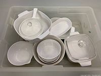 View of various white ceramic CorningWare cookware pieces including a round dish with a glass lid, oval dish, and several bowls inside a container