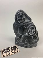 Front view of dark gray soapstone carving depicting two Inuit figures in traditional hooded clothing, showing facial details and original tags for authenticity.