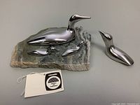 Front and top view of mama loon aluminum sculpture with two babies on natural stone base; separate aluminum duck figurine with tag showing Canadian seal and authenticity.