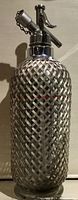 Full view of the antique seltzer bottle showing woven metal mesh body and metal siphon top