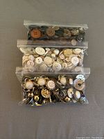 Three sealed plastic bags containing different assortments of buttons: dark buttons, light/neutral buttons with pearl-like finish, and gold-toned decorative buttons.