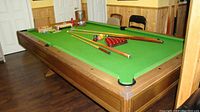 Wide angle photo showing the entire pool table with green felt, wooden frame, pockets and various cues and accessories on the table.