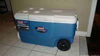 Side view of blue and white Coleman Xtreme cooler showing wheels, handle, lid with cup holders and side handles.