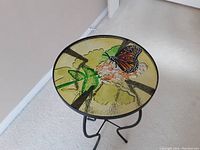 Photo of a round patio table with a textured glass top painted with a colorful monarch butterfly and green leaves. The table has black metal legs and stands about 21 inches tall.