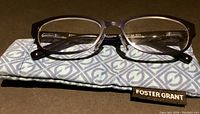 Photo of Foster Grant reading glasses lying on a soft pouch; glasses have brown gradient rectangular plastic frame and clear lenses.