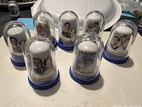 Eight decorative thimbles with images of British royals inside plastic domes with blue bases, displayed on table