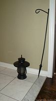 Black metal lantern and curved metal hanging stake shown together on a tiled floor against a beige wall.