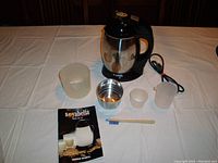 Complete Soyabella SB-130 soy milk maker and grinder with all accessories laid out on a white tablecloth: main unit with handle and stainless steel container, clear measuring cup, small stainless steel bowl, small white plastic cup, cleaning brush, and owner's manual.