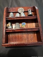 Full view of wooden shelf holding seven assorted thimbles. Visible details include helmet and figure-shaped pewter thimbles, and colorful enamel crests on some thimbles.