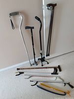 Photo showing six walking canes of various styles, two grabber tools, and two shoe horns leaning against a wall and laid on the floor.