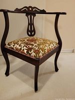 Front angle of vintage corner chair showing dark wood frame, curved arms, intricate carved backrest, and patterned upholstered seat.