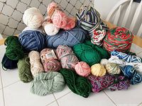 Various cotton yarn skeins displayed on a tiled table and white chair. Colors include greens, blues, cream, pinks, and multicolor patterns.