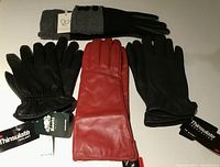 Four pairs of women's gloves displayed together on a flat surface, showing black Roots nappa leather gloves with tags, red leather gloves, black Thinsulate gloves, and black/grey wool gloves with button closures.