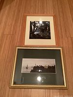 Two framed photographs laid flat on wood floor, one image square with natural wood frame and cream mat, second panoramic in gold frame with dark green mat.