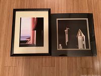 Two framed photographs by S. Lauder displayed side by side on a wooden floor. One is a framed coastal window view photo and the other a matted lighthouse photo.