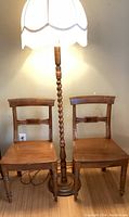 Two wooden chairs with carved rectangular backrests and a wooden spiral floor lamp with a white fabric fringed shade lit up.