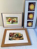 Wide photo showing three framed food-themed art pieces grouped on a surface, including the pear watercolor print, the Peelings photo print, and the Eggsactly vertical triptych.