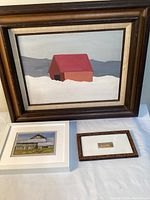 Three framed paintings displayed flat on a white surface: largest is a barn in snow on wood, medium is a barn oil painting, smallest is a tiny cat print.