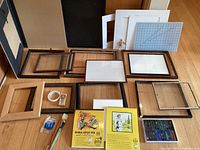 Overview of multiple wooden and plastic picture frames, sketchpads including Manga Artist Pad and pastel set arranged on a wooden floor.