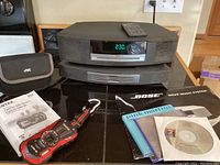 Bose Wave Music System stereo with remote, Pentax Adventure digital camera, JVC case, and manuals on countertop