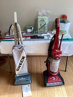 Photo showing Oreck XL Series vacuum alongside red Hoover WindTunnel upright vacuum, mop handles, and various cleaning supplies on a table in background.