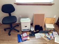 Black fabric swivel office chair with wheels, white Royal shredder, cork board, assorted office supplies including greeting cards, binders, and paper organizers.