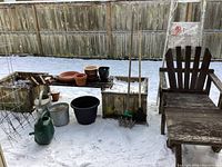 Shows rustic wooden chair with footstool and various garden items on a long bench including pots and watering cans, in a snowy yard.