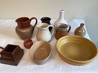 Overview of all pottery items arranged on white cloth, showing pitchers, bowls, square dish and small pots in various colors and styles.